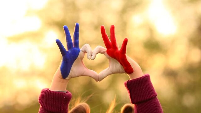 Love and happiness concept. Cute child forming heart gesture with hands outdoors on nature sunset bokeh background. Heart shape of kids hand painted in france flag colors, kids body language