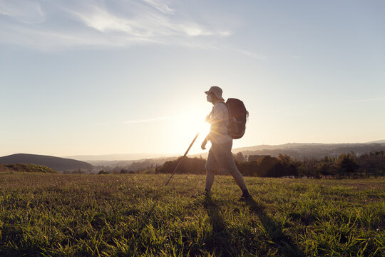 Side view of unrecognizable male pilgrim with rucksack and trekking stick strolling on green meadow under cloudy sky in sunshine in evening in back lit