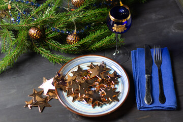 Christmas still life with blue plate with golden stars, wine glass, cutlery, decorated fir tree branches on black background