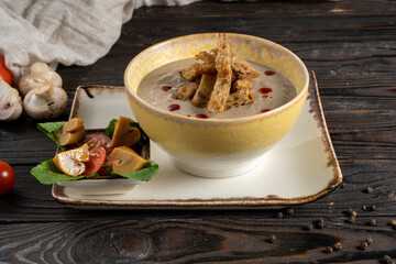 Creamy mushroom soup with rye croutons and pickled champignons. Hot soup in a ceramic bowl on a wooden kitchen table.