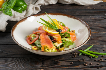 Salad with salmon or trout, bell peppers, olives and olives, garnished with onions and lemon. Cold appetizer of salad leaves and red fish in a ceramic plate on a wooden kitchen table.