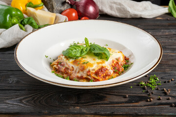 Lasagna with ground beef or minced pork and cheese, garnished with basil leaves and herbs. A hot main course of pasta, meat, cheese and herbs in a ceramic bowl on a wooden kitchen table.