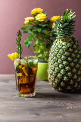 Pineapple tropical cocktail with ice, rum, cola, mint, rosemary  in glass on brown wooden background with yellow roses 