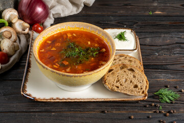 Solyanka. Traditional Russian soup made from vegetables, bacon and herbs in a ceramic plate with sour cream and rye bread on a wooden kitchen table. Hodgepodge