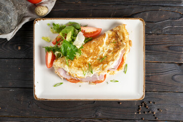 Omelet with ham, bacon, cheese and tomatoes. Classic hot dish of eggs, milk, meat, cheese, vegetables and salad in a ceramic plate on a wooden kitchen table.