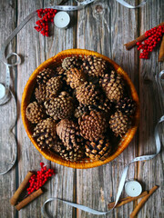 Orange wicker basket with pine cones shot from above wooden table with white ribbon, tealights, cinnamon sticks and red berries around it. Top view, flat lay. Winter and Christmas background.