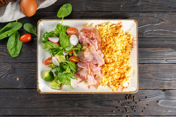 Scrambled omelet with bacon and vegetable salad. A hot breakfast dish of eggs, milk, meat, lettuce, radish, cucumber and tomato in a ceramic plate on a wooden kitchen table.