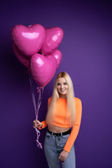 Happy blonde with heart-shaped balloons on a purple background in the studio.