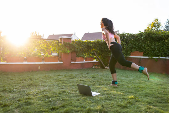 Side view of fit female athlete in sportswear using laptop and doing exercise while following online tutorial during training in backyard at sunset