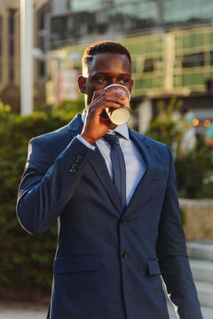 Serious adult formally dressed African American male manager drinking takeaway coffee while standing against blurred urban background of modern city