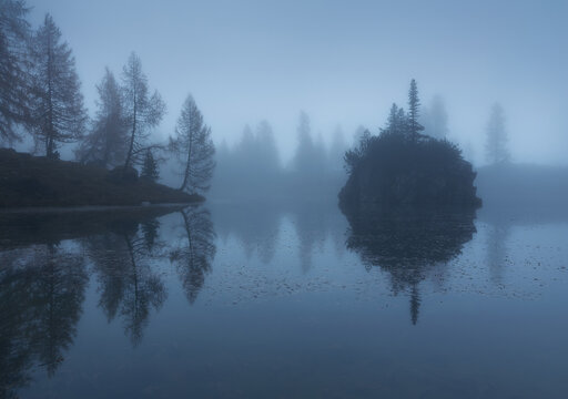 Scenic view of river with rocky formation near hill with growing trees in mist under blue sky in winter