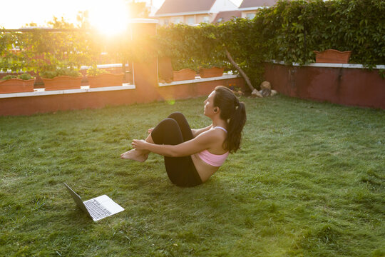 Side view of fit female athlete listening to online tutorial on laptop and doing abdominal crunches while balancing on grass during training at sunset