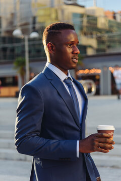 Serious adult formally dressed African American male manager drinking takeaway coffee while standing against blurred urban background of modern city