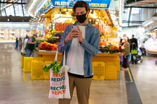 Young male buyer in protective mask holding reusable cotton bag with groceries and browsing smartphone during shopping in grocery market