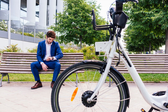 Handsome businessman in elegant clothes sitting on wooden bench while checking time near electric bike in town