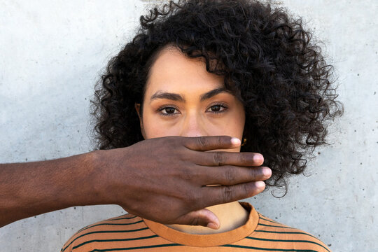 Anonymous African American male friend covering face of young attentive female with Afro hairstyle near cement wall while looking at camera