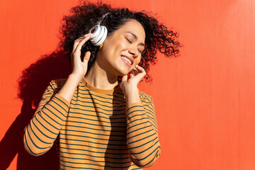Happy trendy ethnic female with closed eyes and Afro hairstyle enjoying song in headset on red background
