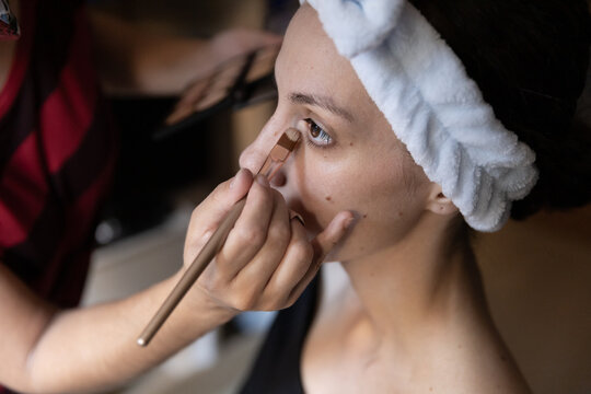 Cropped unrecognizable professional stylist applying concealer on female customer lower lid by using brush in studio