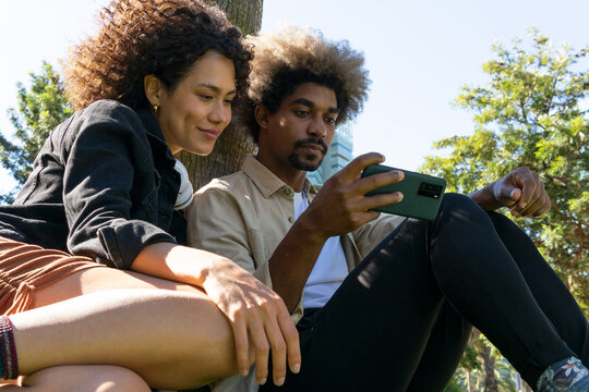 Low angle of positive young African American millennials in trendy outfits watching video on smartphone while sitting on grass in park