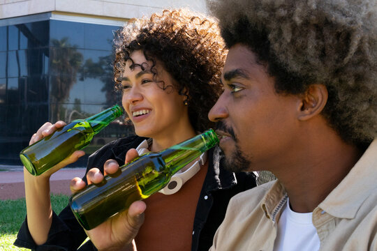 Side view of positive young African American millennial in casual clothes drinking beer while spending time together in street cafe