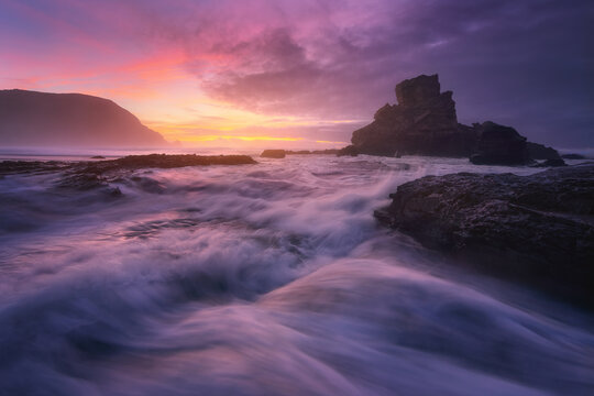 Rocky formations on rippled ocean with fast water flow under cloudy sky with glowing sun at night