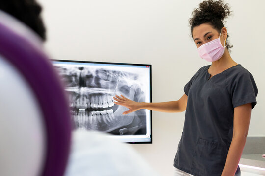 Friendly African American female dentist in uniform and medical mask examining X ray of jaws and teeth while sharing information with unrecognizable patient