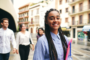 Young smiling woman with Afro braids and plastic folder looking at camera behind multiracial coworkers