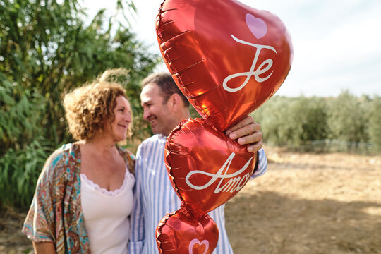Tender mature couple standing together in garden with red air balloons in shape of heart and looking at each other during stroll
