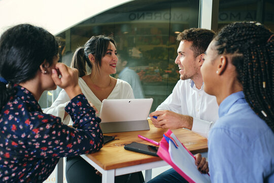 Cheerful multiethnic colleagues talking while looking at each other and sitting at table with netbook in building