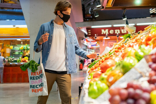 Young male customer in protective mask carrying eco friendly recycled fabric shopping bag and choosing fresh groceries while buying food in supermarket