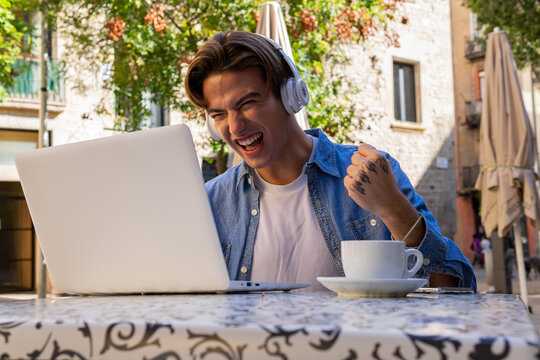 Overjoyed young male in wireless headphones getting incredible news on laptop and celebrating victory while sitting at table with cup of coffee on cafe terrace