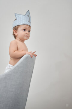 Cheerful little boy wearing toy crown standing in armchair and clapping hands while looking away