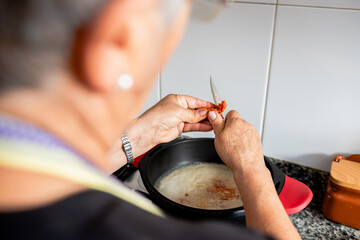 Back view of crop housewife adding red pepper to pan with sauce while preparing traditional Catalan cod dish in kitchen
