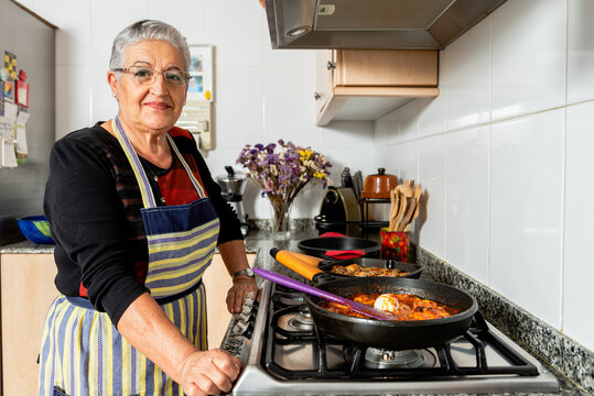 Senior housewife cooking fish steaks in frying pan while preparing typical Catalan cod dish in kitchen looking at camera