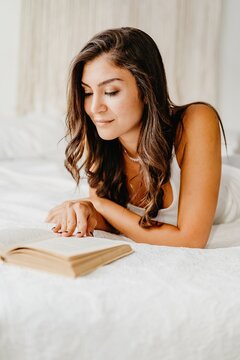 Positive young female in underwear leaning on cozy bed and reading book while spending weekend morning in light bedroom with boho decoration