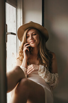 Young cheerful female in stylish wear talking on cellphone while looking away near window at home