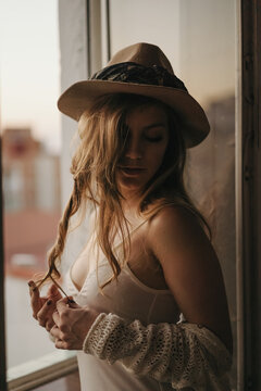 Young seductive female in stylish apparel and straw hat standing with closed eyes near window in apartment