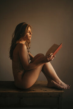 Side view of sensual female wearing lace underwear sitting on wooden table with book and enjoying story