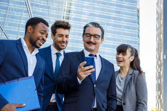 Group of happy multiracial colleagues in classic suites laughing while reading good news on smartphone standing together on street