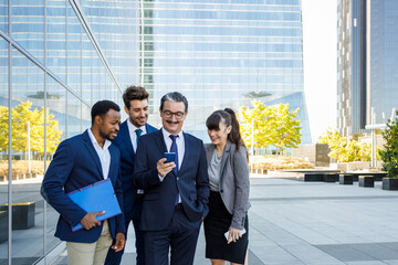 Group of happy multiracial colleagues in classic suites laughing while reading good news on smartphone standing together on street
