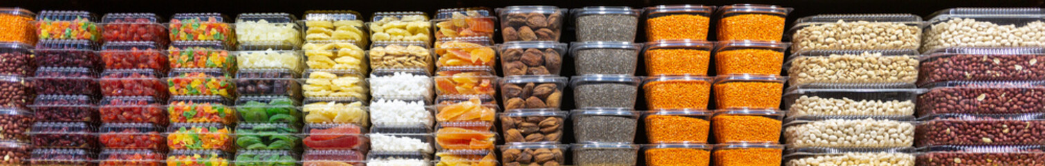 Many plastic boxes with different nuts, pieces of coconut, dried pieces of bananas, kiwi, cranberries, lentils, figs, dried apricots as a background, texture, pattern. Horizontal banner for websites.
