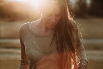 Crop serene female wearing transparent sweater standing in lush green park in evening sunlight and looking down pensively