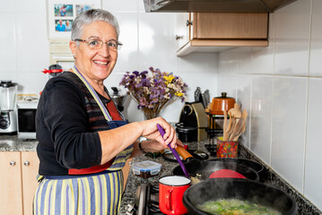 Happy mature woman frying red onion in hot pan with oil while preparing traditional Catalan fish dish in home kitchen looking at camera