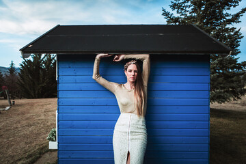 Positive calm female wearing stylish outfit and hair wreath leaning on rural plank construction in countryside