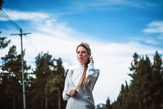 Charming young female in white casual outfit and hairband touching hair gently while standing near coniferous woods and looking at camera
