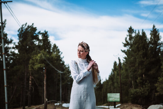 Charming young female in white casual outfit and hairband touching hair gently while standing near coniferous woods and looking at camera