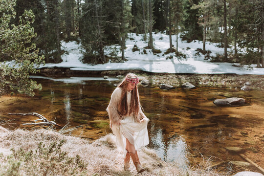 Side view of charming young long haired female in Nordic style boho clothes and wreath strolling along river shore in sunny early spring day with snowy forest landscape in background