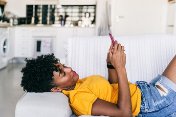 Side view of excited African American millennial female in casual outfit browsing mobile phone and laughing while resting on sofa in modern apartment