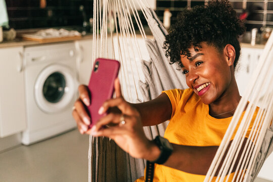 Positive African American curly haired female student in casual clothes sitting in hammock and taking selfie on mobile phone while spending free time at home