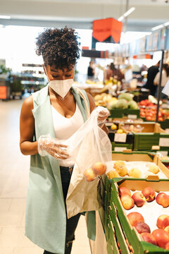 Side view of African American female customer in protective mask and gloves picking fruit from box while making purchases in supermarket during coronavirus pandemic